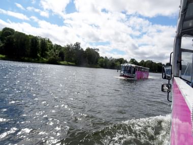 Blick von einem rosa-weißen Ausflugsboot auf einer Stockholmer Wasserstraße, mit einem weiteren Boot voraus und grünem Ufer unter bewölktem Himmel.
