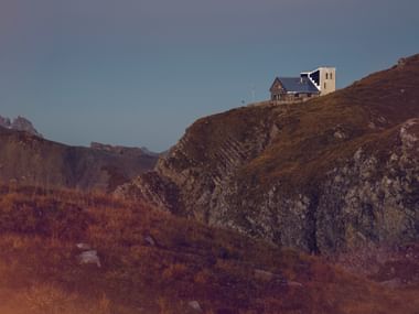 Refuge Rambert Hütte perché sur une crête alpine rocheuse avec végétation automnale et sommets montagneux en arrière-plan.
