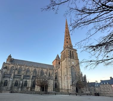 Gotische Kathedrale St-Tugdual in Tréguier mit hohem Turm und Steinfassade unter blauem Himmel, eingerahmt von kahlen Ästen auf einem Platz.
