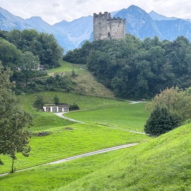 Steinturm auf bewaldetem Hügel, umgeben von grünen Wiesen und Wegen. Alpenberge erheben sich im Hintergrund unter bewölktem Himmel.