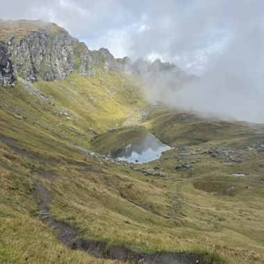 Kleiner Bergsee in einem grasbewachsenen Tal mit felsigen Gipfeln und Nebel. Ein Wanderweg schlängelt sich durch die Alpenlandschaft.
