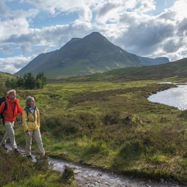 Zwei Wanderer in bunten Jacken gehen auf schlammigem Pfad durch schottische Highlands mit Bergen, Bach und weißem Cottage im Hintergrund.