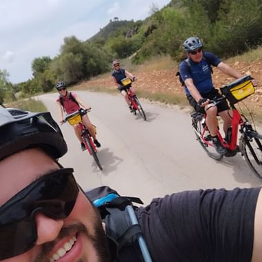 Ein Mann mit Velohelm und Sonnenbrille macht ein Selfie beim Velofahren. Im Hintergrund folgen vier weitere Personen mit Helm und Sonnenbrille auf rotem Velo.