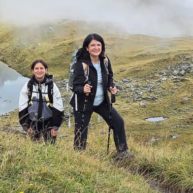 Zwei Wanderinnen mit Rucksäcken und Wanderstöcken auf einer nebligen Almwiese in der Ostschweiz. Felsige Hänge und Nebel im Hintergrund.