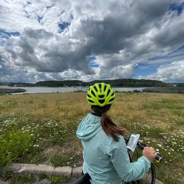 Eine Fahrradfahrerin mit hellblauer Jacke und gelb-gruenem Helm blickt über eine Wiese mit weissen und gelben Blumen. Im Hintergrund ist ein See und ein bewoelkter Himmel.
