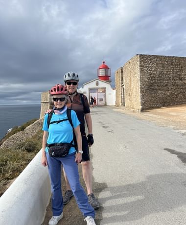 Zwei Radfahrer mit Helmen und Sonnenbrillen am Leuchtturm Cabo de São Vicente in der Algarve. Leuchtturm mit roter Kuppel und Steinfestung.