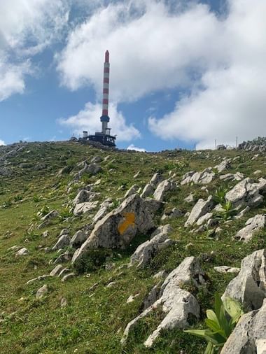 Rot-weiß gestreifter Funkturm auf einem felsigen Hang im Jura mit Kalksteinfelsen, grüner Vegetation und bewölktem Himmel.
