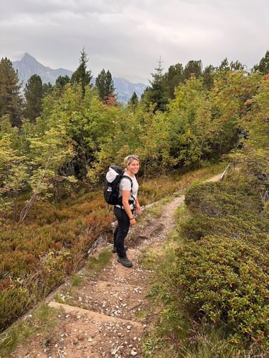 Wanderin Sara mit Rucksack auf Bergweg bei Gurnigelpass, umgeben von herbstlich gefärbten Bäumen und Bergen im Hintergrund.