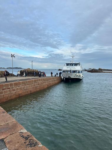 Weiße Fähre mit Aufschrift Bréhat nähert sich einem Steinpier in der Bretagne. Passagiere warten am Kai unter bewölktem Himmel mit Felsküste.