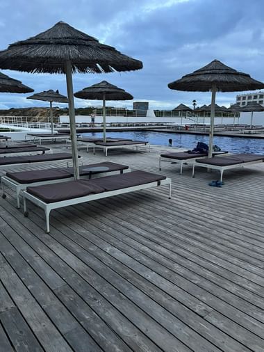 Holzpooldeck mit weißen Liegen und Strohdachschirmen im Hotel Memmo Baleeira in Sagres unter bewölktem Abendhimmel.