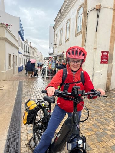 Radfahrerin in roter Regenjacke und Helm auf E-Bike in nasser Kopfsteinpflasterstraße in Albufeira. Fußgänger mit Schirmen im Hintergrund.