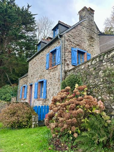 Traditionelles bretonisches Steinhaus in Tréguier mit hellblauen Fensterläden und Schornstein, umgeben von Hortensien und grünem Garten mit Steinmauer.