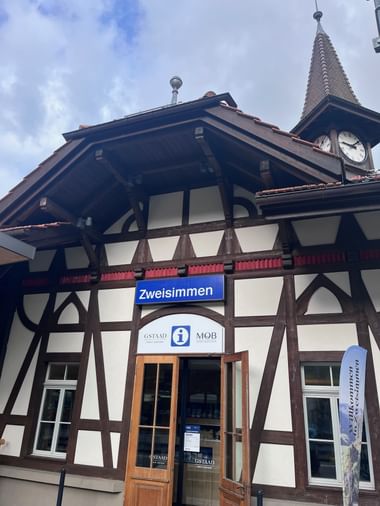 Traditionelles Schweizer Fachwerkgebäude mit Bahnhof Zweisimmen und Uhrturm. Blaues Bahnhofsschild und Touristeninformation sichtbar.