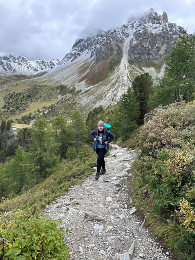Wanderin mit Rucksack auf steinigem Weg am Alpenpässeweg. Schneebedeckte Berggipfel und grüne Alpenvegetation im Hintergrund sichtbar.