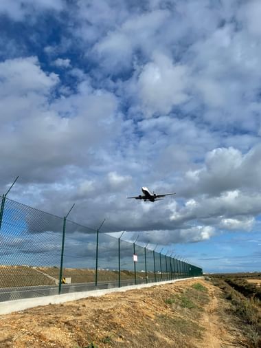 Flugzeug steigt über dem Flughafen Faro auf, mit grünem Sicherheitszaun entlang eines Feldwegs unter bewölktem Himmel in der Algarve.