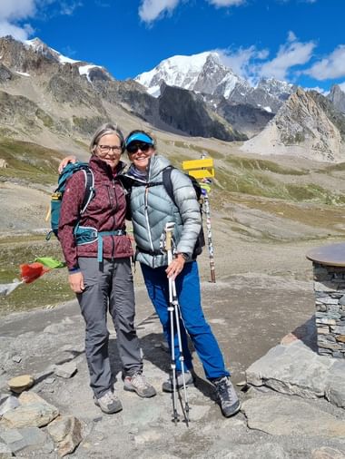 Zwei lächelnde Wanderer mit Rucksäcken und Trekkingstöcken im Vallée des Glaciers. Schneebedeckte Mont-Blanc-Gipfel unter blauem Himmel.