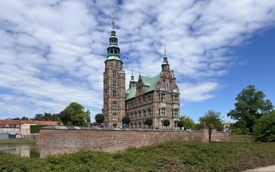 Schloss Rosenborg mit roten Backsteinmauern und grünen Kupfertürmen in Kopenhagen, umgeben von einem Wassergraben und grünen Rasenflächen.