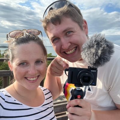 Eine Frau und ein Mann sind draussen und lachen in eine Kamera. Der Mann hält eine Filmkamera mit Mikrofon in der Hand.