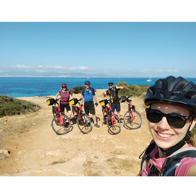 Eine Frau mit Velohelm und Sonnenbrille macht ein Selfie. Im Hintergrund sind drei Personen, ebenfalls mit Helm und vier Velos. Dahinter ist das Mittelmeer.