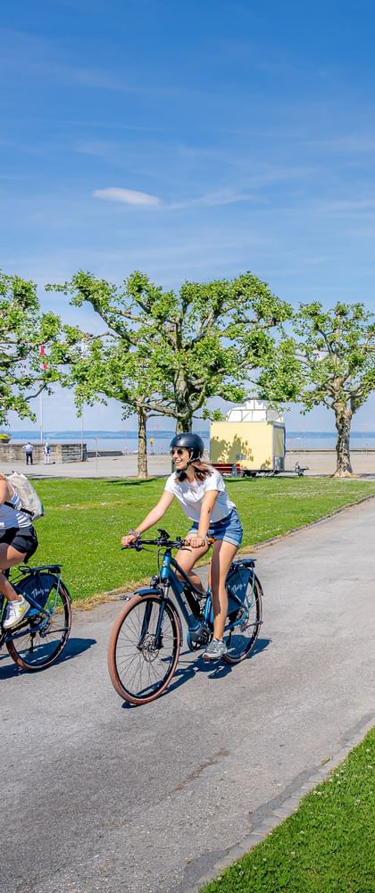 Zwei Radfahrer auf einem Uferweg in Arbon mit grünen Rasenflächen, Bäumen und dem Bodensee im Hintergrund unter blauem Himmel.