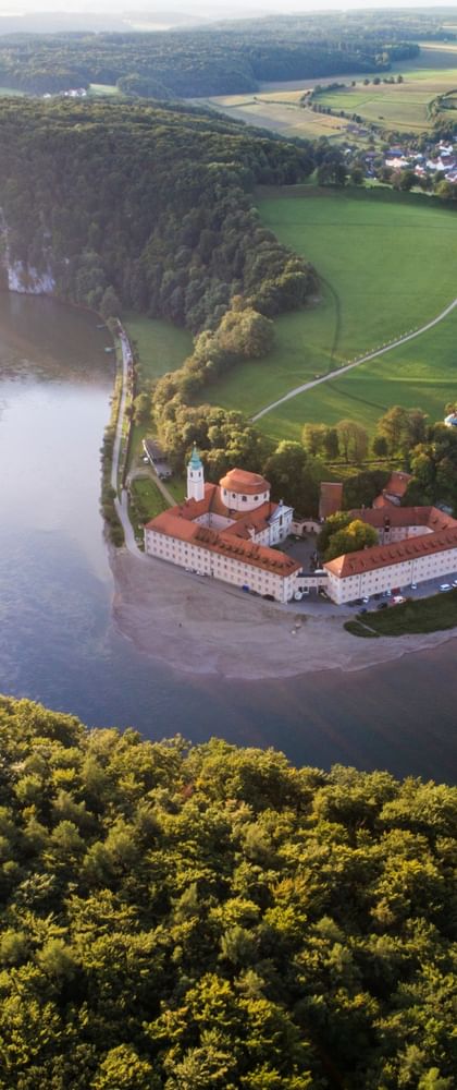 Luftaufnahme von Kloster Weltenburg auf einer Halbinsel, umgeben von der Donau. Dichte Wälder und grüne Felder erstrecken sich über die Landschaft.