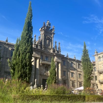 Steinkathedrale mit barocken Türmen und verzierter Fassade, flankiert von hohen Zypressen, umgeben von angelegten Gärten unter blauem Himmel.