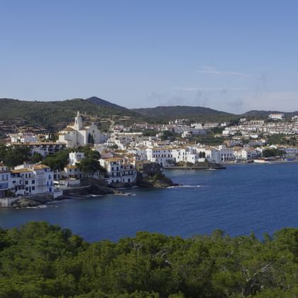 Panoramablick auf Cadaqués, ein weißes Küstendorf an einer Bucht mit blauem Wasser, umgeben von grünen Hügeln und Bergen unter klarem Himmel.