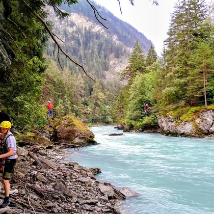 Menschen mit Sicherheitsausrüstung überqueren einen türkisfarbenen Fluss an Seilen im Kletterpark Pfunds, Österreich, umgeben von Wald.