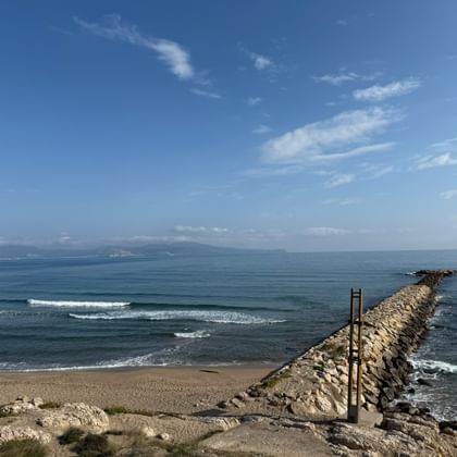 Steinerner Wellenbrecher erstreckt sich ins ruhige blaue Meer mit Sandstrand im Vordergrund. Berge am fernen Horizont unter klarem Himmel.