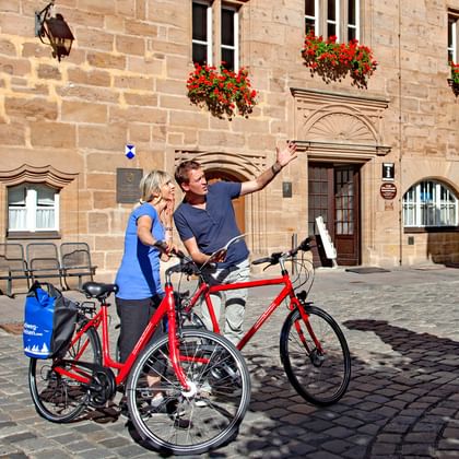Zwei Radfahrer machen ein Selfie mit roten Tourenrädern auf Kopfsteinpflaster in Ansbach, umgeben von historischen Sandsteingebäuden mit Blumenkästen.