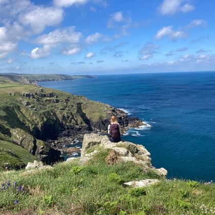 Person sitzt auf felsiger Klippe mit Blick auf den blauen Atlantik und die dramatische Küste Cornwalls mit grünen Feldern und Klippen.