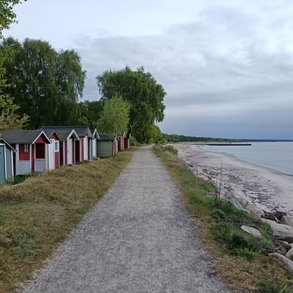 Schotterweg entlang der Küste mit bunten Strandhütten links und Sandstrand rechts unter bewölktem Himmel am Österlenleden.