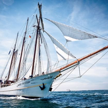 Weißes Segelschiff Mare Frisium mit türkisfarbenem Rumpf segelt auf blauem Wasser mit weißen Segeln vor bewölktem Himmel.