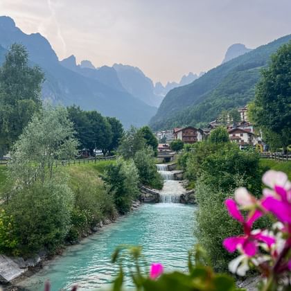 Türkisfarbener Rio Masso Bach mit Wasserfall fließt durch Alpendorf, rosa Blumen im Vordergrund, traditionelle Gebäude und Berge dahinter.