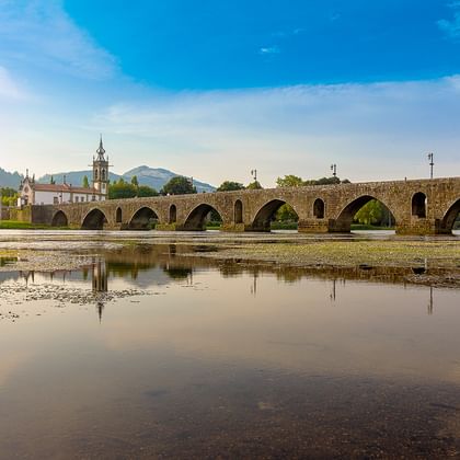 Mittelalterliche Steinbrücke mit mehreren Bögen über einen Fluss in Ponte de Lima, Portugal. Kirchturm und Hügel im Hintergrund sichtbar.
