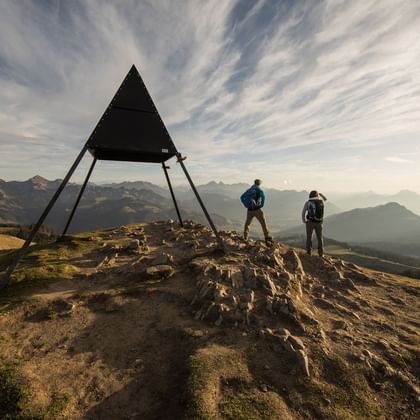 Zwei Wanderer stehen auf einer Bergspitze und blicken in die Berglandschaft vor Ihnen. Neben den Wanderern steht ein Metalliges Dreieck dessen Spitz sich in den Himmel richtet.