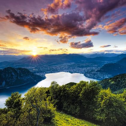 Coucher de soleil spectaculaire sur le lac de Lugano au Tessin avec des nuages colorés, des montagnes et une forêt verte luxuriante au premier plan.
