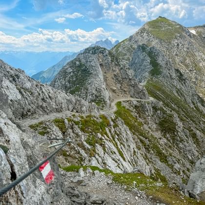 Rot-weiße Wegmarkierung am Nordkette Berggrat mit felsigen Gipfeln und gewundenem Pfad. Alpine Landschaft zwischen Garmisch und Bozen sichtbar.