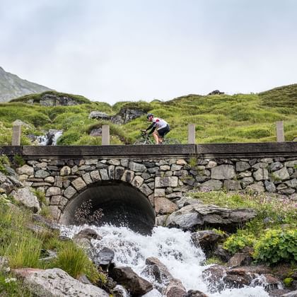 Ein Biker fährt über eine steinerne Brücke bei der aus einem Bogen das Wasser durchfliesst.