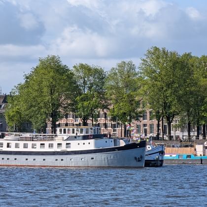 Großes grau-blaues Hausboot Floor van Aemstel am Amsterdamer Kanal mit historischen Gebäuden und grünen Bäumen am Ufer.