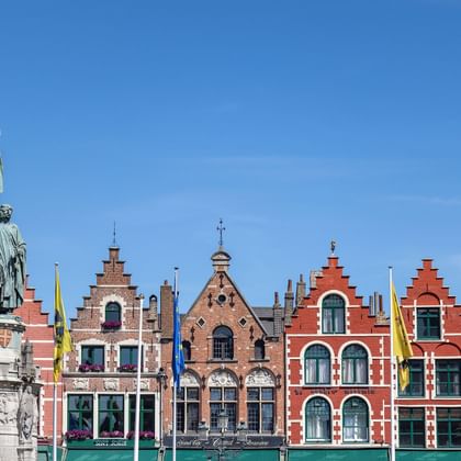Bronzedenkmal auf Steinsockel vor bunten mittelalterlichen flämischen Gebäuden mit Treppengiebeln am Marktplatz von Brügge.