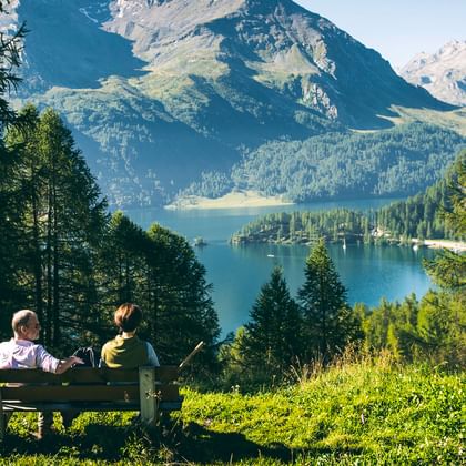 Zwei Wanderer machen eine Pause auf einem Bank in der Wiese. Vor ihnen der Ausblick auf eine romantische Berglandschaft mit See.