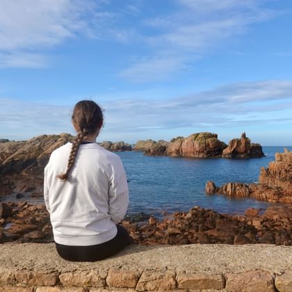 Person mit Zopf sitzt auf Steinmauer mit Blick auf rosa Granitfelsen und blaues Meer auf der Île de Bréhat, Bretagne.