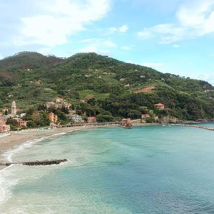 Luftaufnahme eines Cinque Terre Küstendorfs mit bunten Häusern entlang eines geschwungenen Strands, umgeben von grünen Hügeln und türkisem Meer.