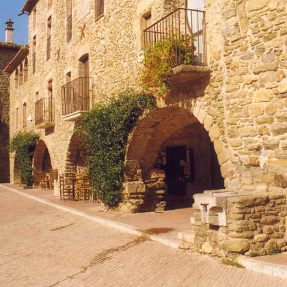 Mittelalterliche Steingebäude mit gewölbten Türen und Balkonen in einem nordkatalanischen Dorf. Ein Steinbrunnen steht im sonnigen Innenhof.
