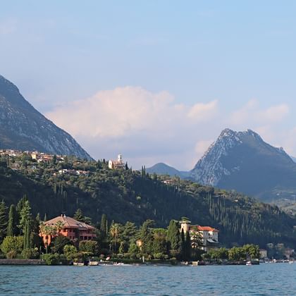 Lac de Garde avec des villas élégantes le long du rivage, entouré de collines boisées et de pics alpins spectaculaires en arrière-plan sous un ciel bleu.