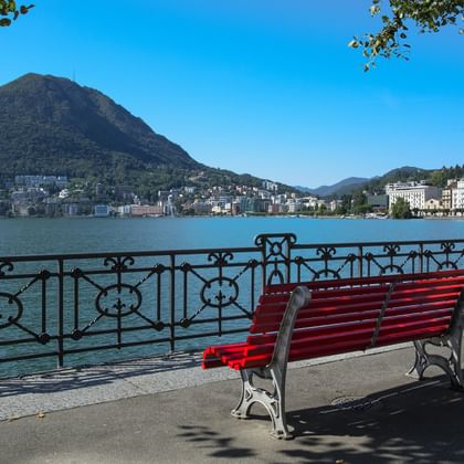Rote Holzbank an der Seepromenade in Lugano mit verziertem Eisengeländer, Blick auf türkisfarbenen See und Monte San Salvatore.