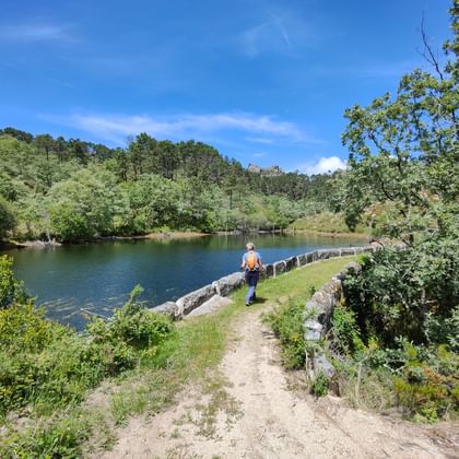 Wanderer mit orangem Rucksack auf schmaler Steinmauer über einem See im Peneda-Gerês-Nationalpark, umgeben von grünen Wäldern unter blauem Himmel.