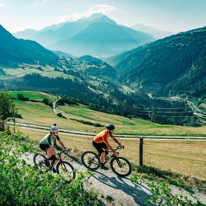 Two cyclists riding gravel bikes on a dirt path through green meadows in Graubünden, with forested mountains and a valley in the background.