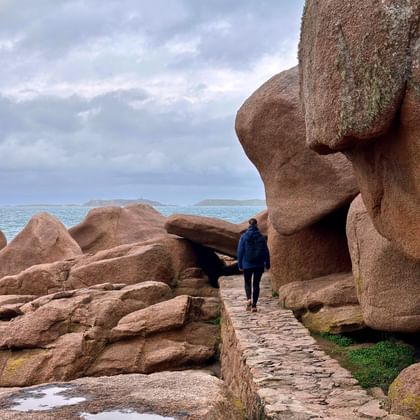 Person in blue jacket walking on stone path between massive pink granite rock formations at Ploumanach coast, Brittany. Cloudy sky and sea in background.